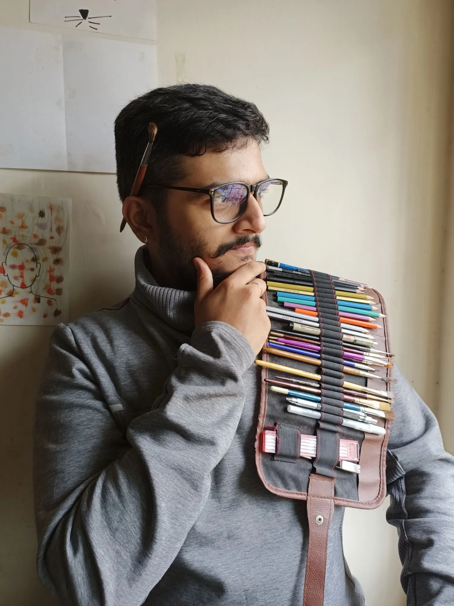 Suhruth in glasses and a grey sweatshirt, a paintbrush tucked behind his ear, chin in hand, with an open roll of pencils and brushes draped over his shoulder.