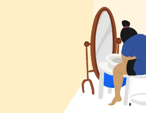A person with their hair in a bun hunched over a pottery wheel beside a tall oval mirror, in a warm cream room.