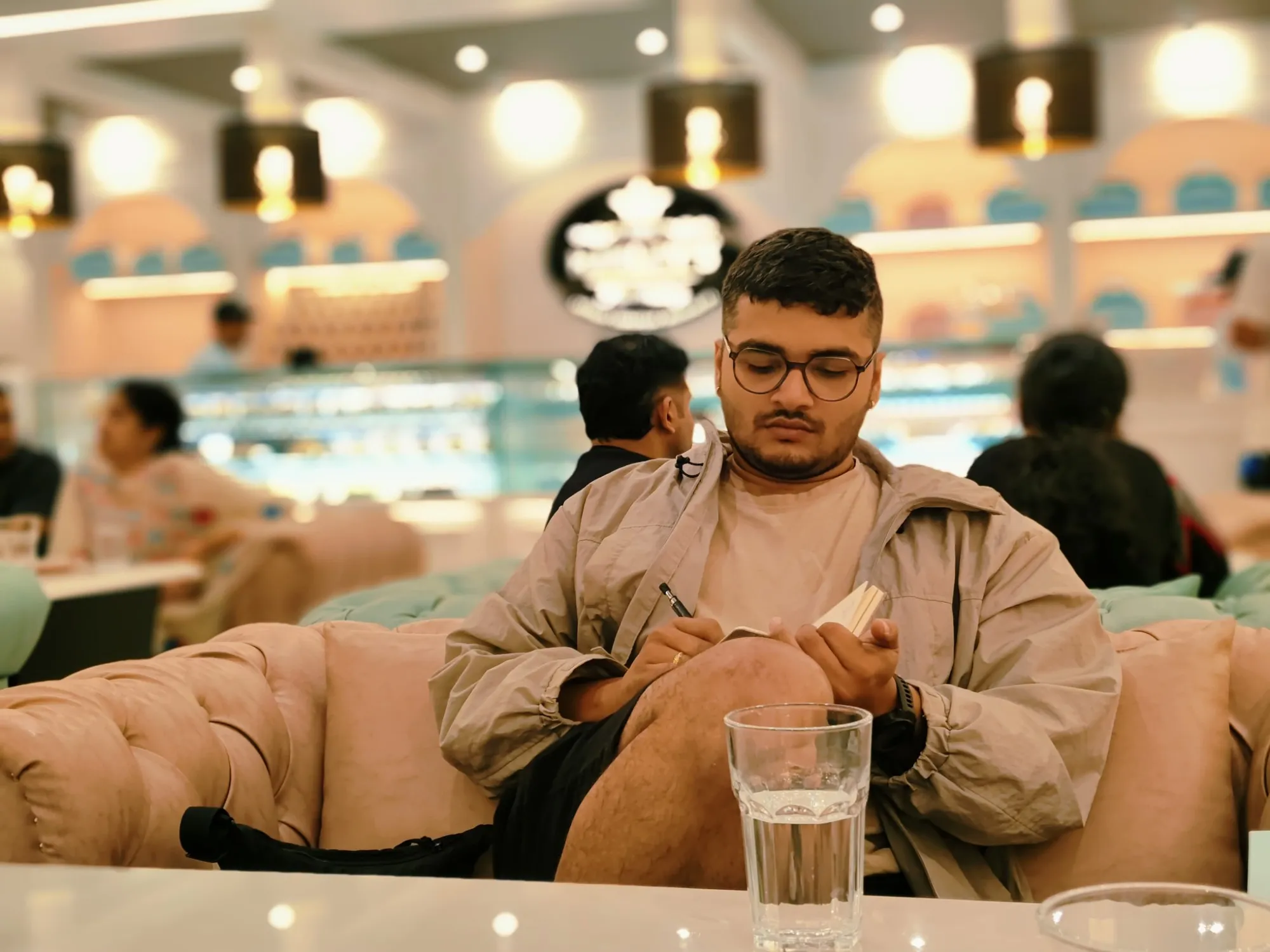 Suhruth in a café, in glasses and a beige windbreaker, sketching in a small notebook with a glass of water on the table in front of him.