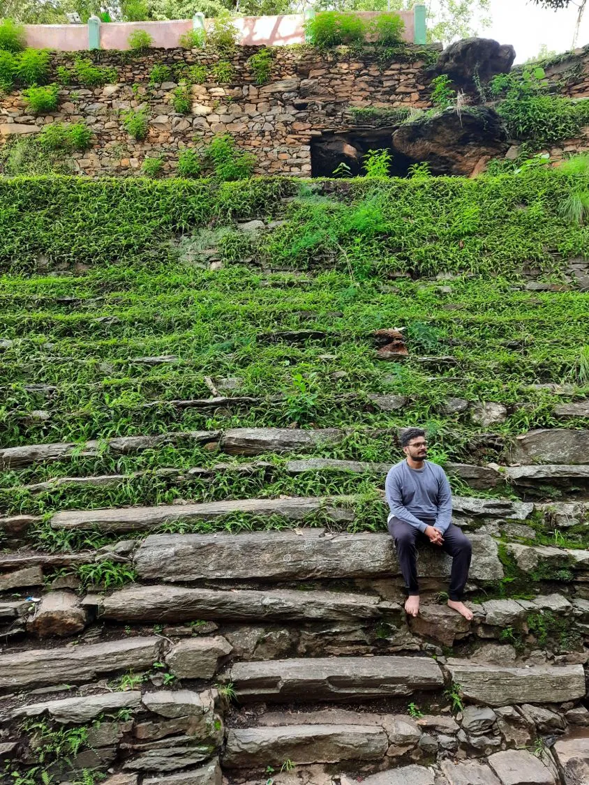Suhruth sitting barefoot on old stone steps overgrown with ferns and grass.
